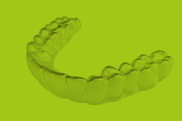 Wir sehen eine Aligner-Schiene auf grünen Hintergrumd, Das Grün ist die CI-Farbe der Zahnarztpraxis. Ein sattes kräftiges leuchtendes Grün mittlerer Helligkeit. Die Aligner-Schiene bildet die Zähne perfekt nach, ist dabei aber so durchsichtig wie man es von Verpackungen und PET kennt. Sie sieht fest und stabil aus. Man kann sich gut vorstellen, dass diese Schiene beim Tragen so gut wie nicht auffällt.